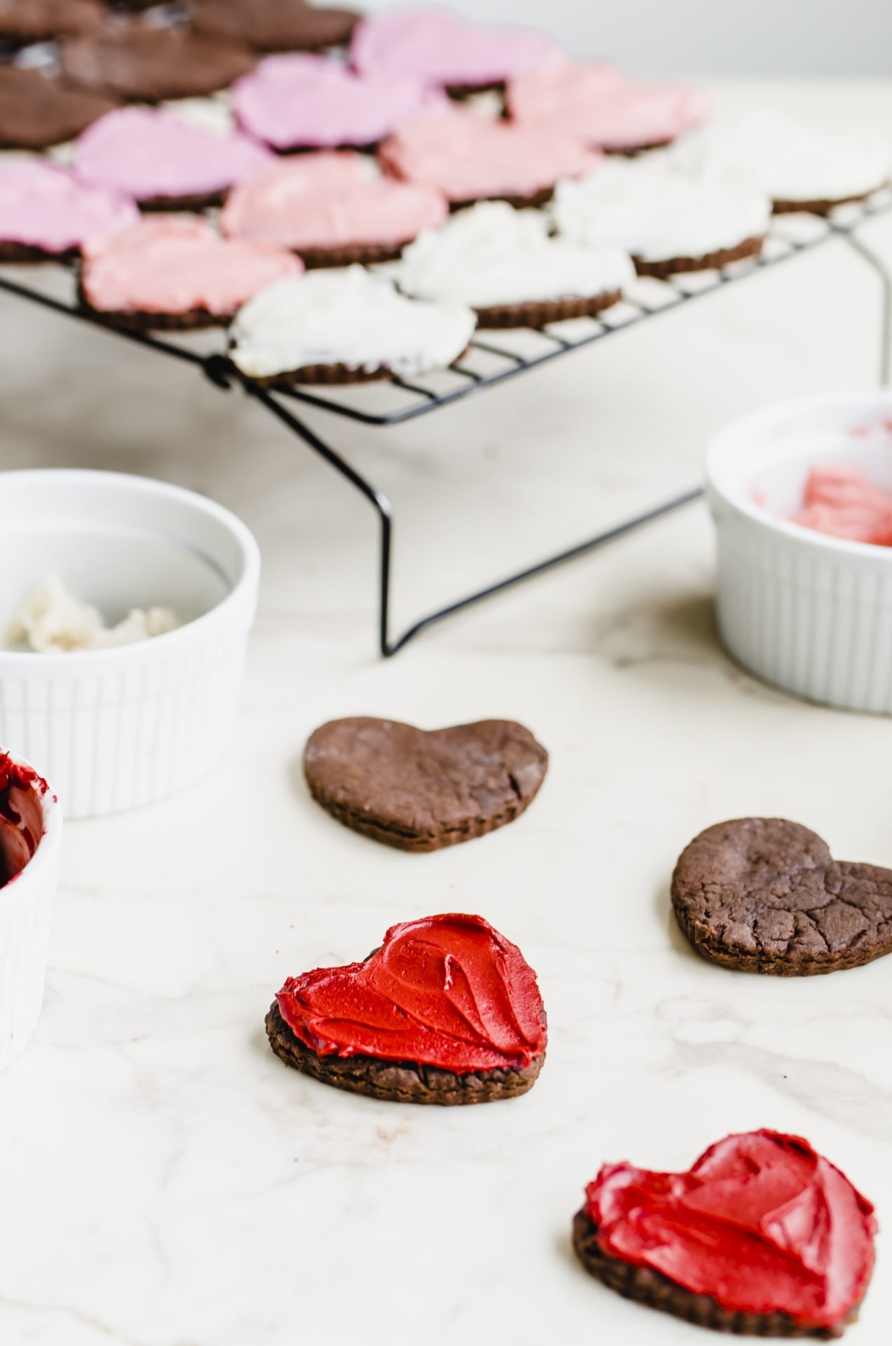 Frosted heart chocolate cutout cookies on a white countertop with bowls of frosting on the side.