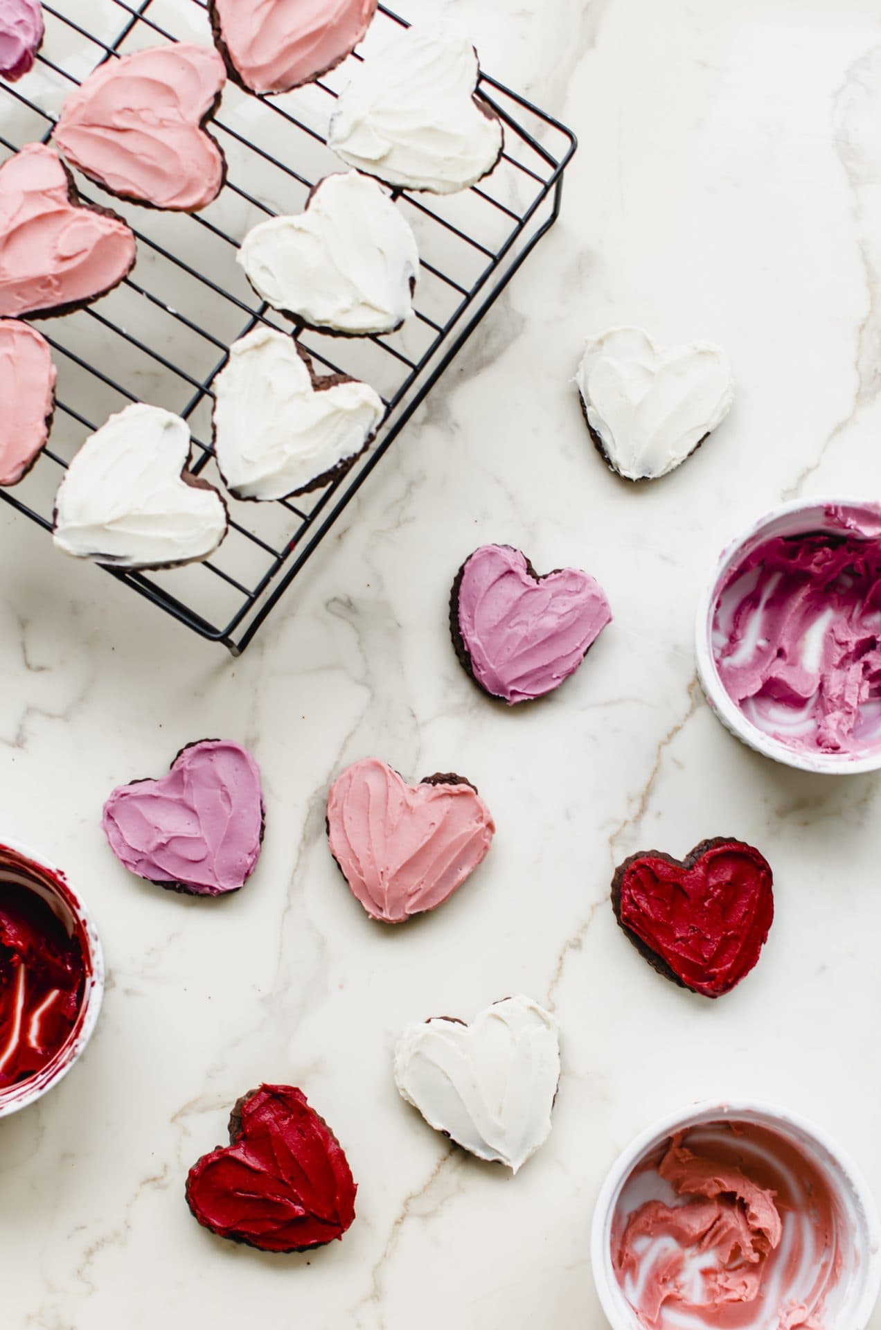 Frosted heart chocolate cutout cookies on a white countertop with bowls of frosting on the side.