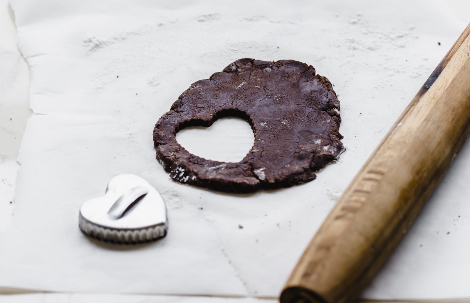 A heart cookie cutter next to rolled out chocolate cookie dough with a rolling pin on the side.
