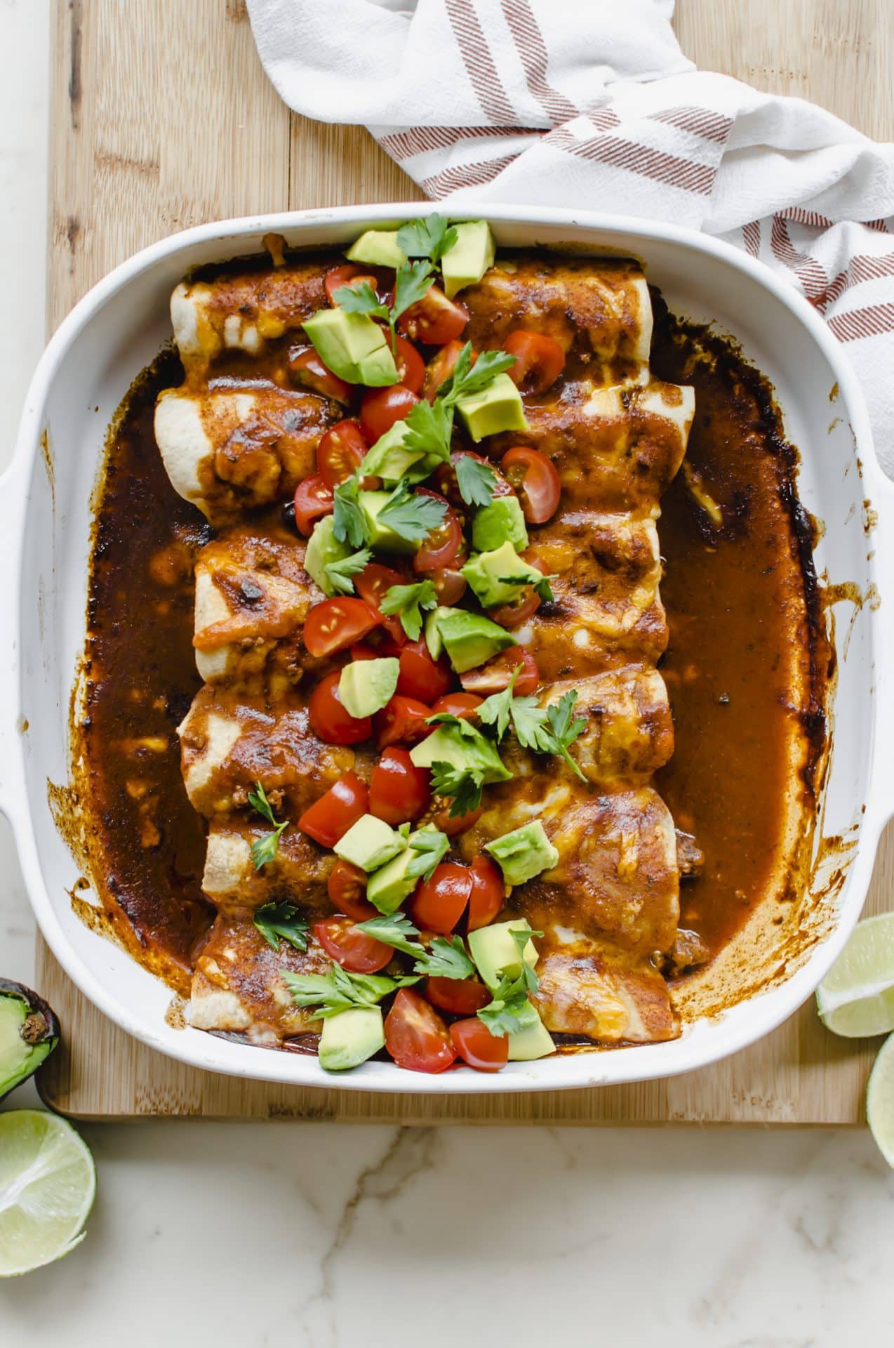 A white baking dish filled with turkey and poblano enchiladas topped with sliced avocado and tomatoes.