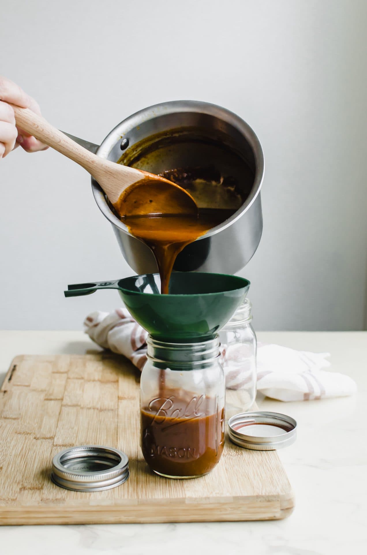 A hand pouring a saucepan of enchilada sauce into a mason jar through a canning funnel.