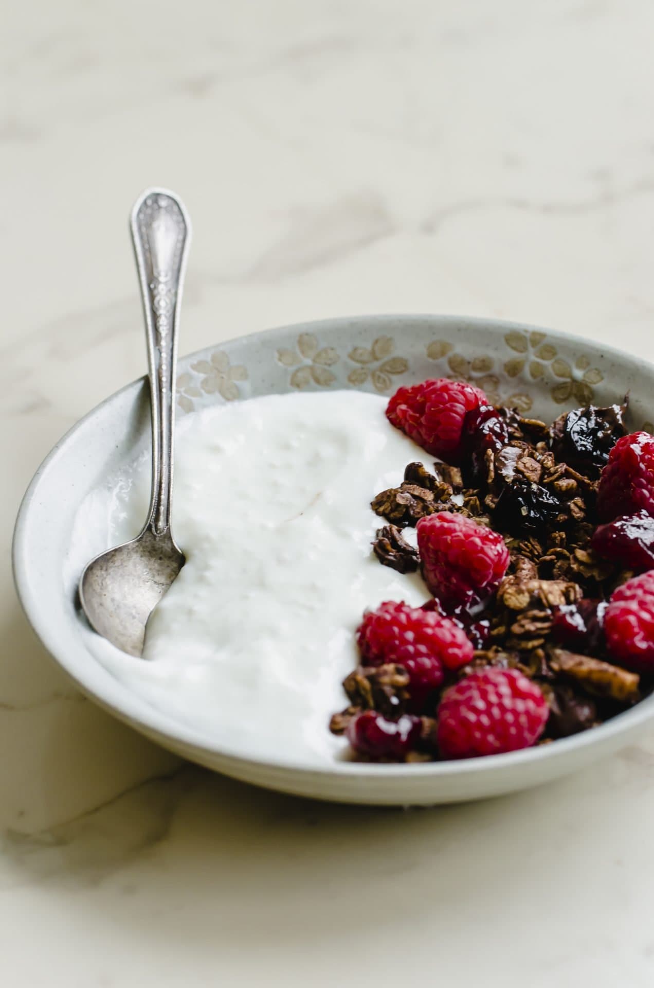 A gray stone bowl of yogurt topped with chocolate granola and raspberries with a spoon on the side.