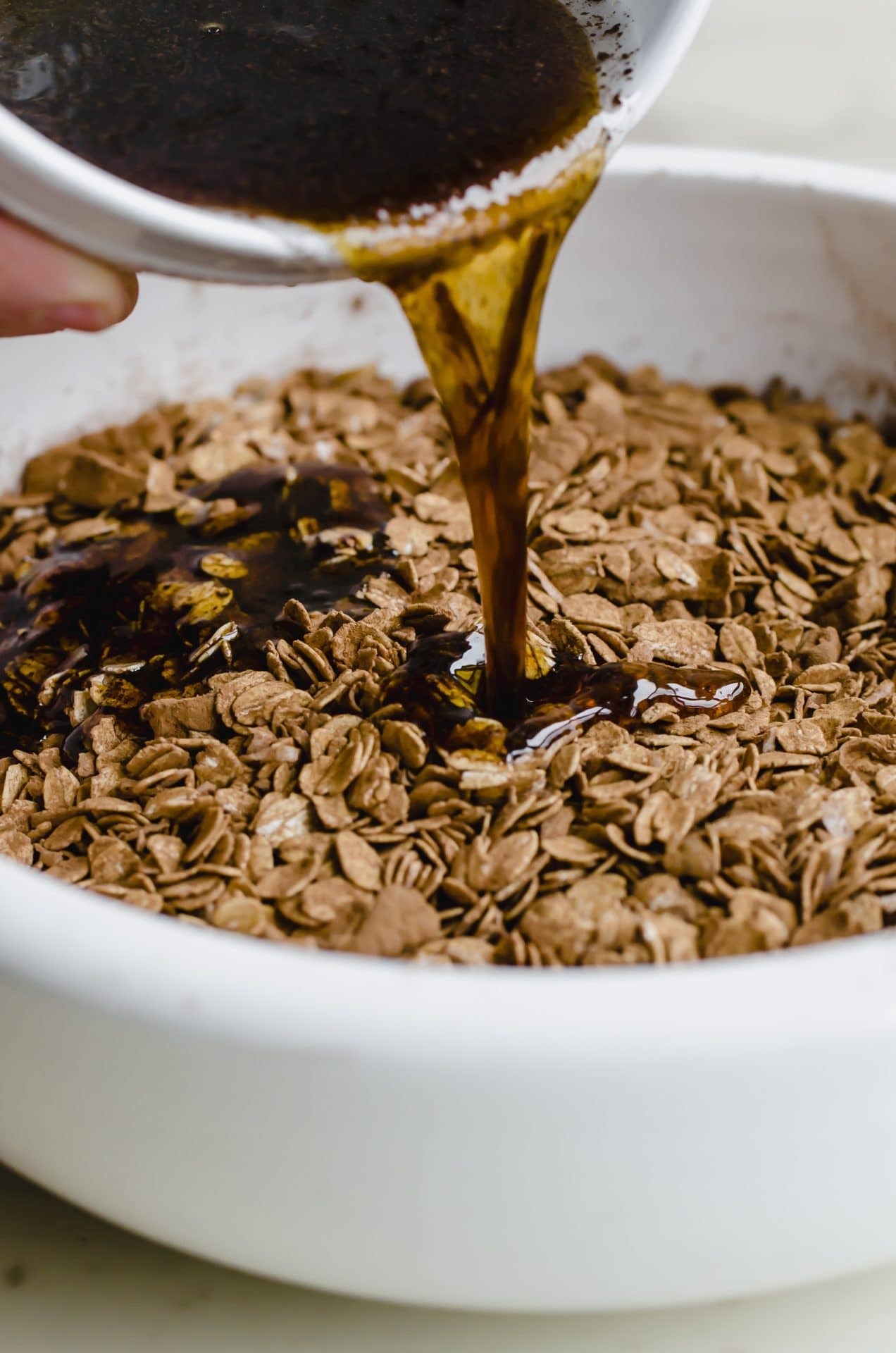 Maple syrup mixture pouring into an oat granola mixture in a white bowl.