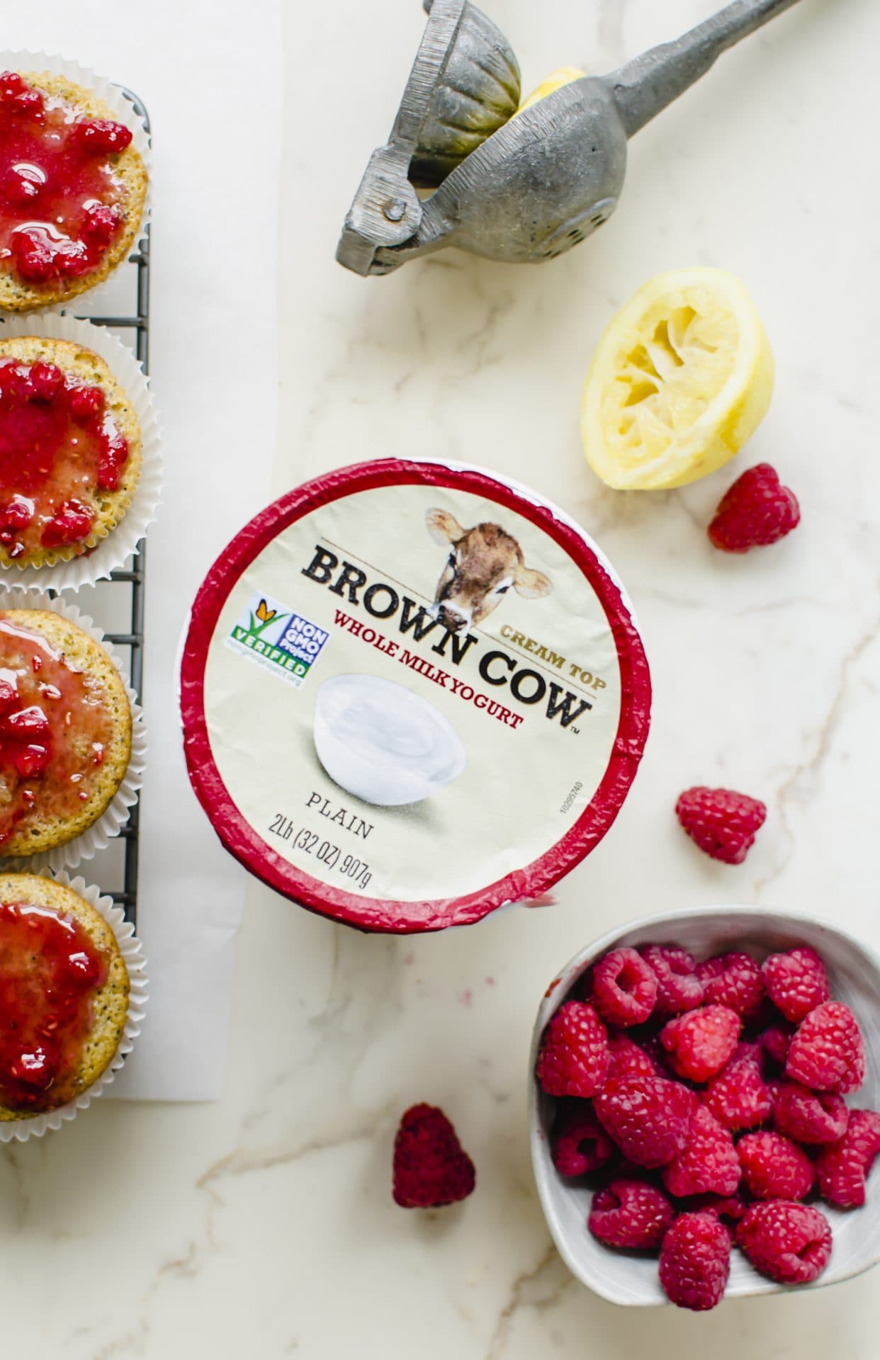 An overhead shot of a container of Brown Cow yogurt with muffins and a bowl of raspberries on the side.
