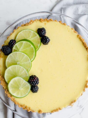 A fresh lime tart with sliced lime and blackberries on top. This is an overhead view and on a white marble background.