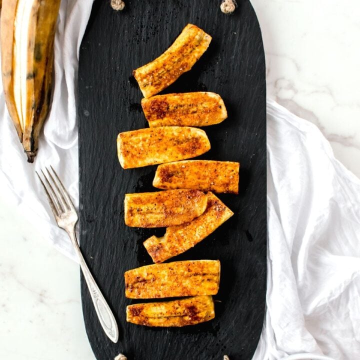 Overhead view of a slate tray with roasted plantains distributed over the top. Cinnamon is sprinkled on top of the plantains.