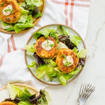 Two salmon cakes with aioli and basil on a bed of lettuce plated with a fork on the side.