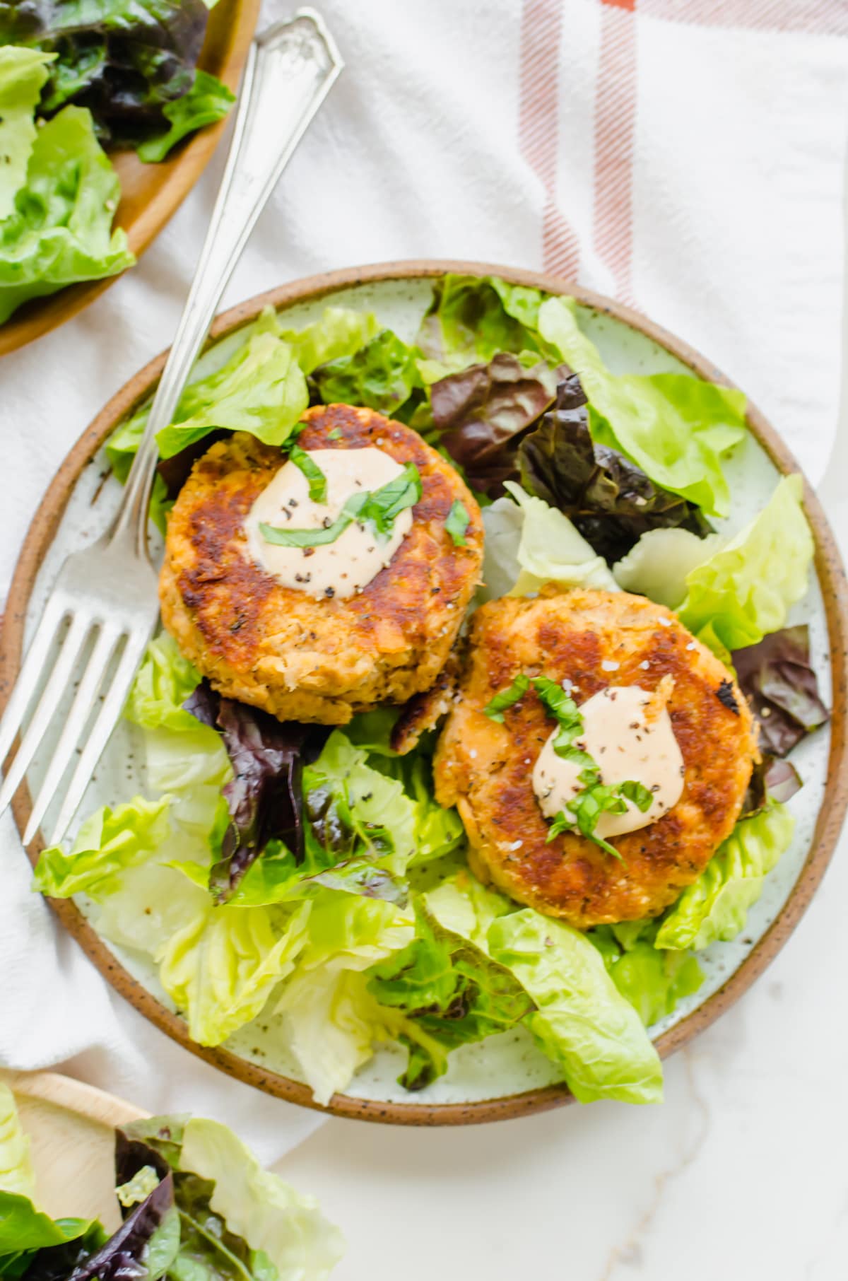 Two salmon cakes with aioli and basil on a bed of lettuce plated with a fork on the side. 