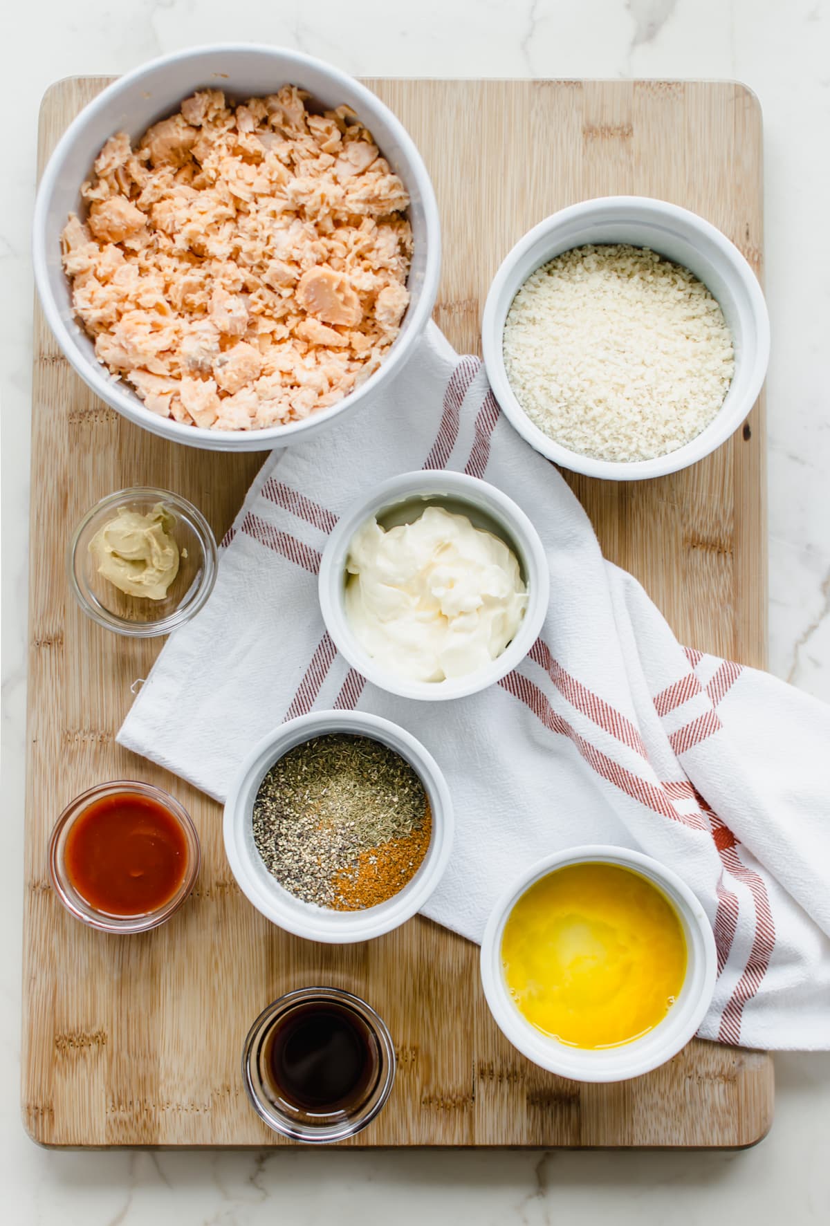 Portioned out ingredients in white bowls on a cutting board for making salmon cakes. 