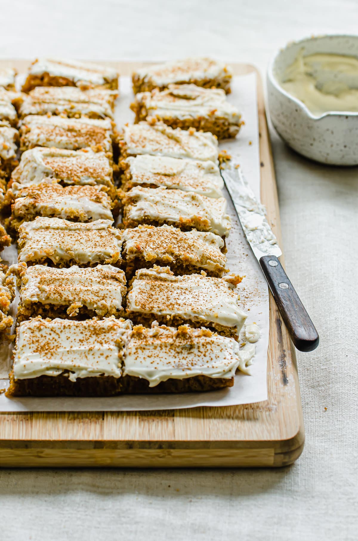 Sliced carrot cake bars on a cutting board with a bowl of frosting in the background.