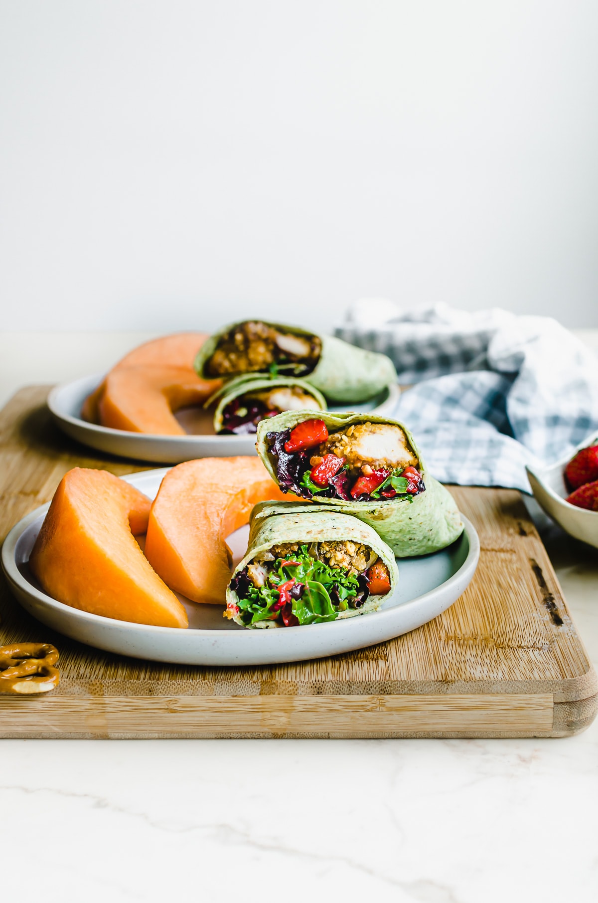 Plates of chicken wraps with pieces of cantaloupe on a cutting board with a gingham dish towel. 