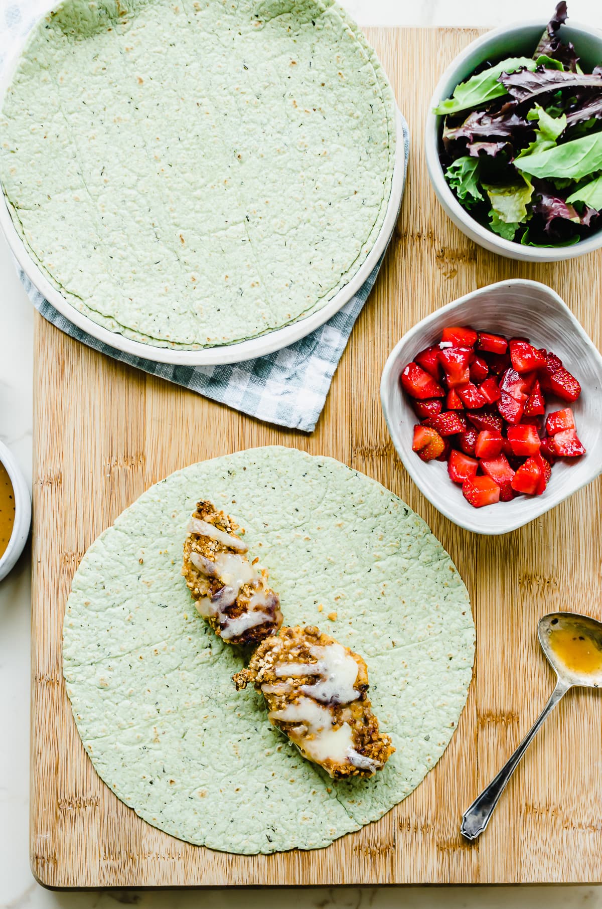 Chicken strips on a spinach wraps with bowls of strawberries and lettuce on the side.