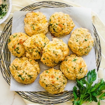 A wire charger lined with parchment with cheddar bay biscuits on top with yellow dish towel and sprigs of fresh parsley.