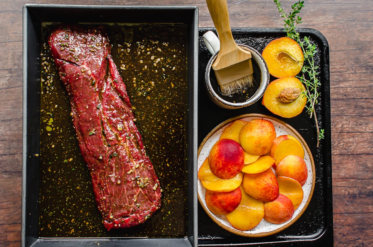 Marinated steak in a pan with a plate of peaches and a cup of basting sauce with a brush.