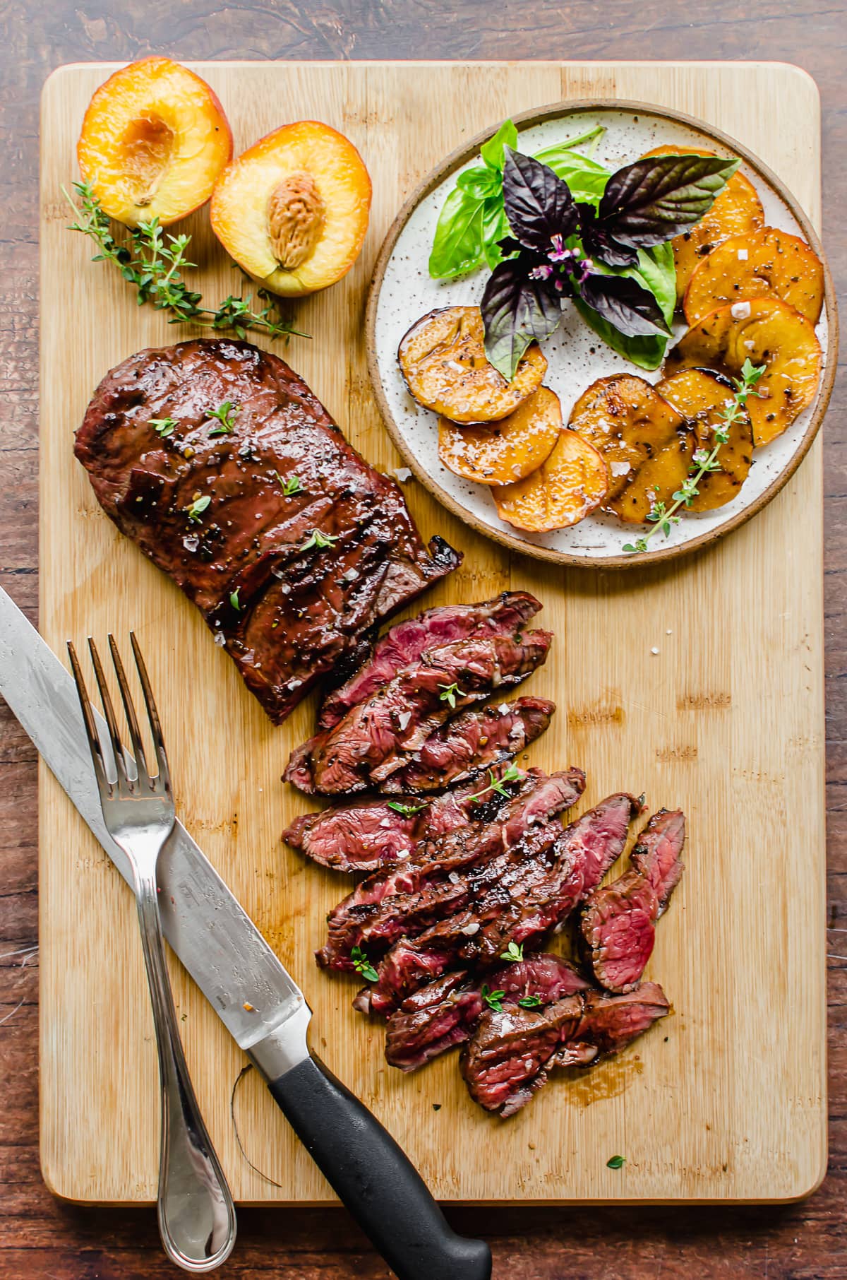 Overhead shot of balsamic steak being sliced on a cutting board with a plate of peaches and basil on the side.