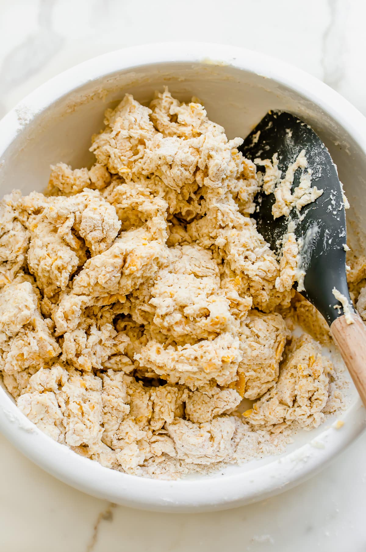 A rubber spatula folding biscuit dough in a white bowl.