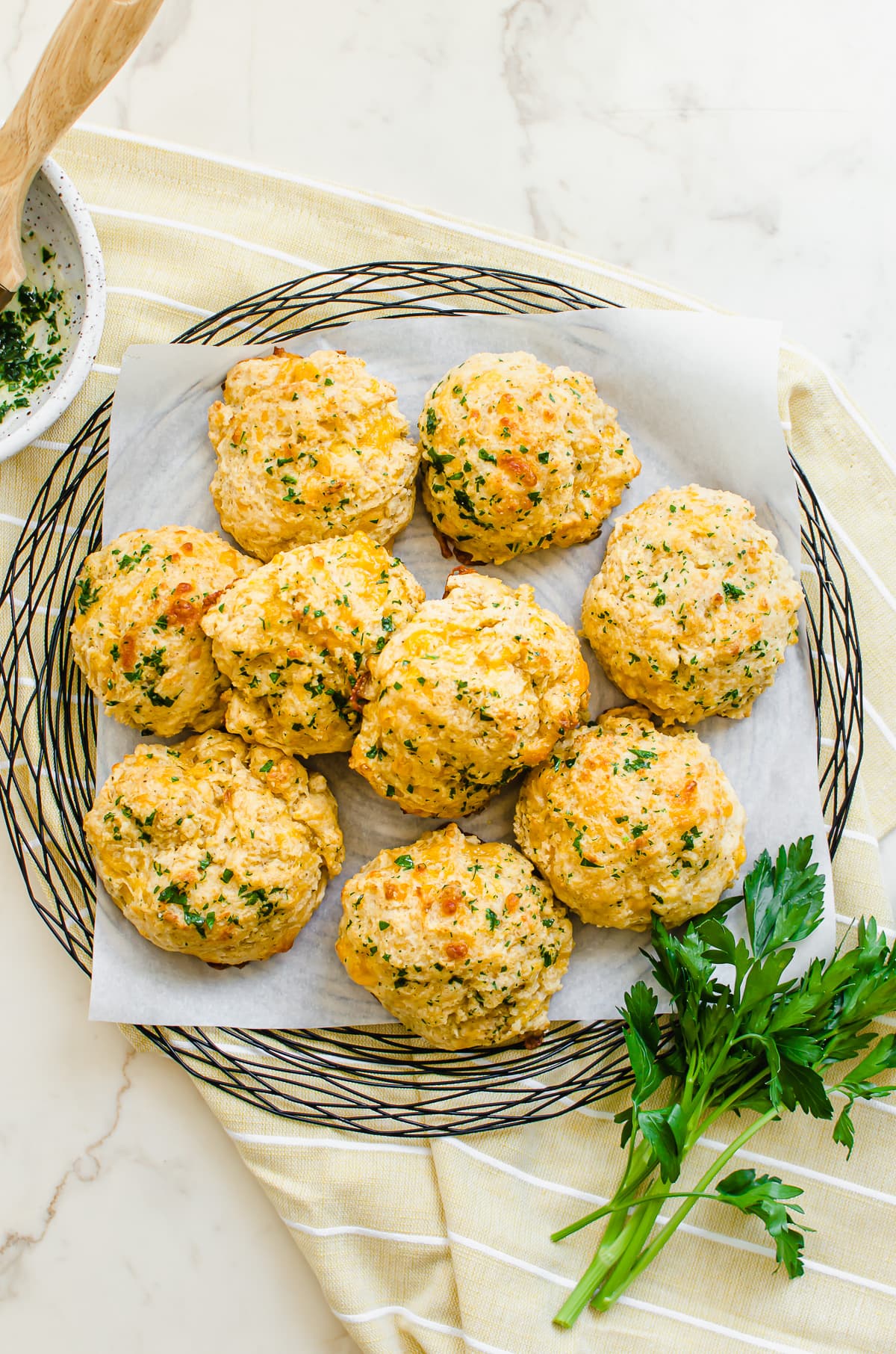 A wire charger lined with parchment with cheddar bay biscuits on top with yellow dish towel and sprigs of fresh parsley.