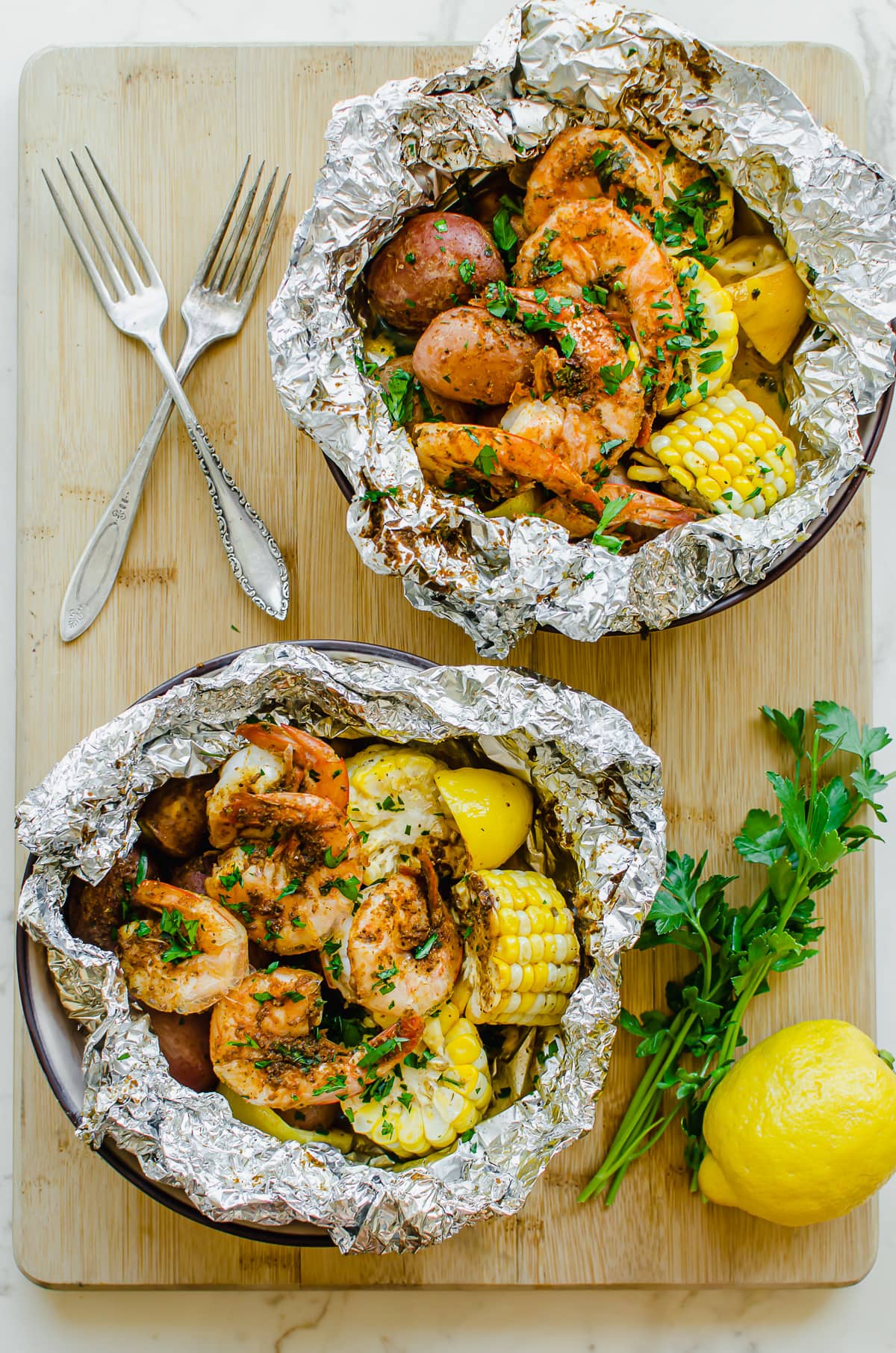 Two bowls of shrimp boil in foil on a wood cutting board with forks on the side.