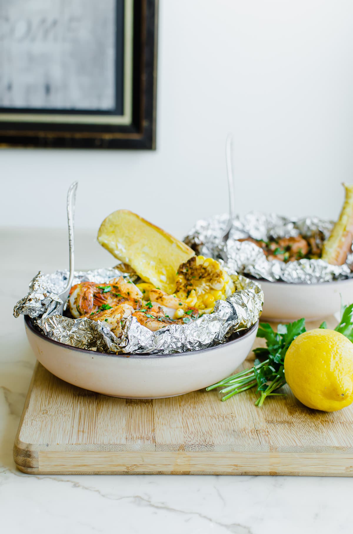 Two bowls of grilled shrimp boil on a wood cutting board with fresh lemon and parsley on the side.