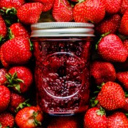 A jar of strawberry jam on top of a bed of fresh strawberries.