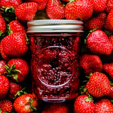 A jar of strawberry jam on top of a bed of fresh strawberries.