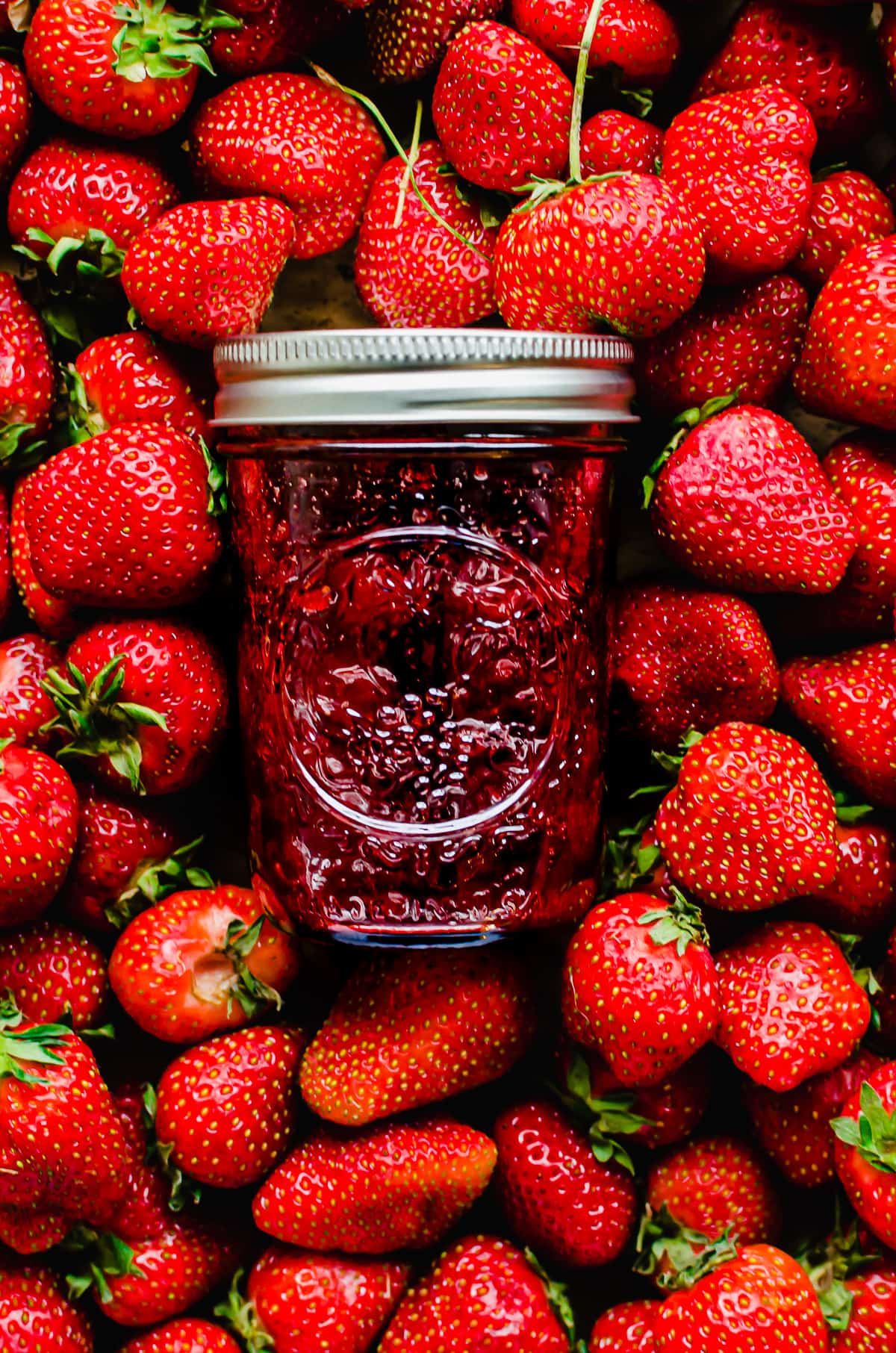 A jar of strawberry jam on top of a bed of fresh strawberries.