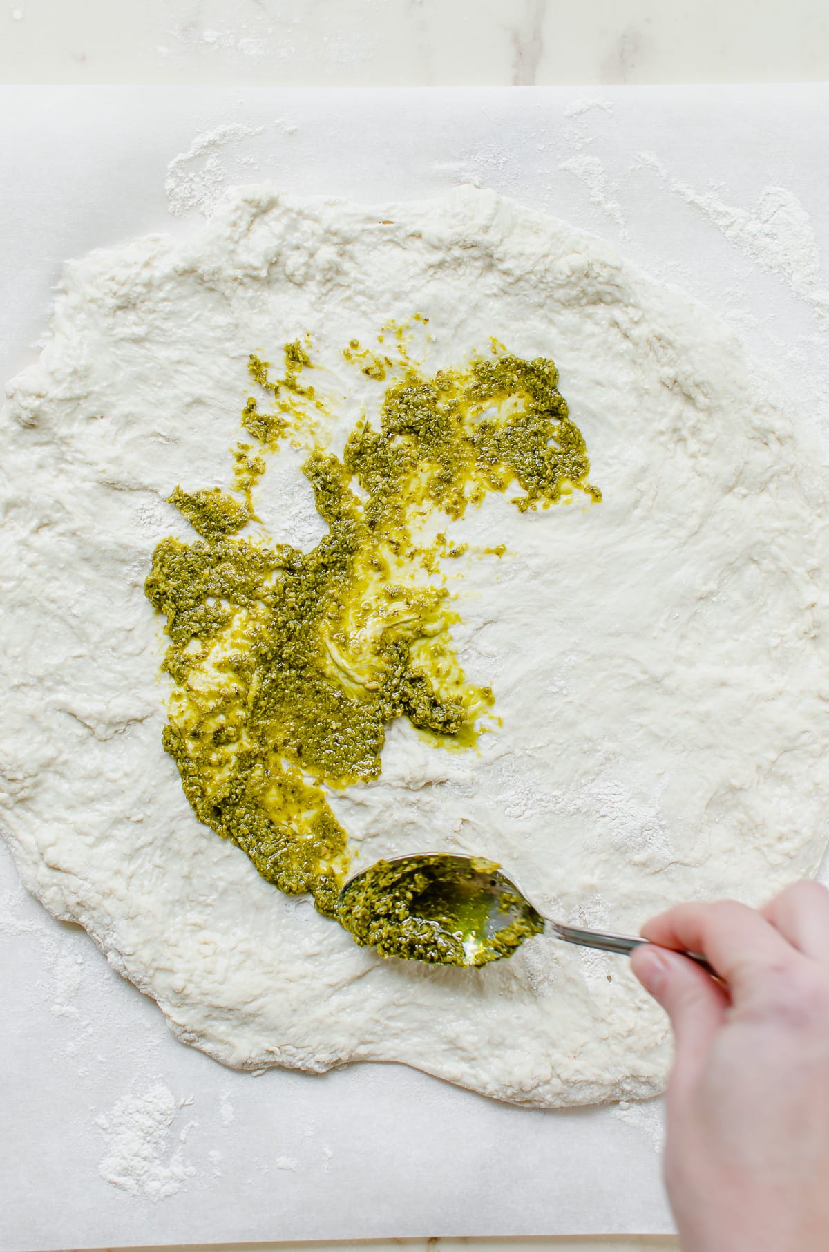 A hand spreading pesto on rolled out pizza dough.