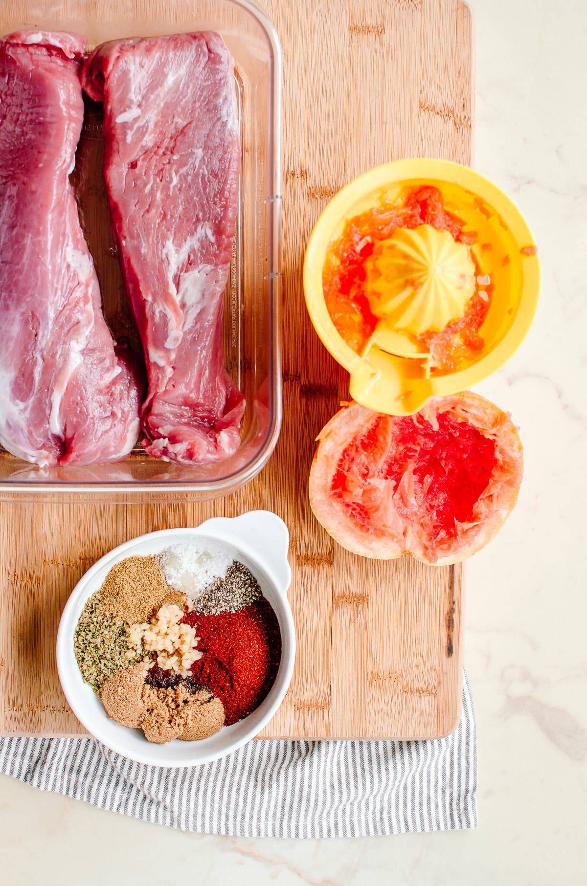 Two pork tenderloins in a clear dish with a bowl of spices for a rub and a juicer with grapefruit on the side.