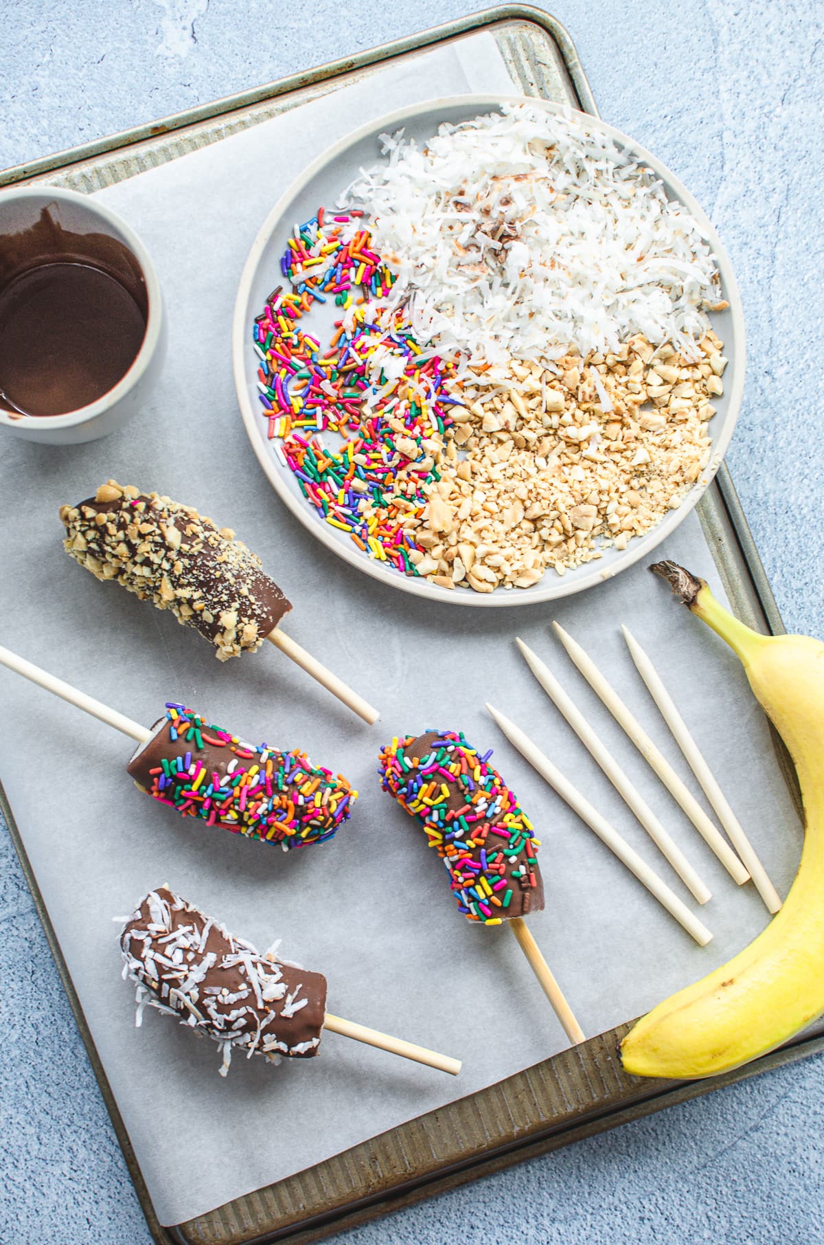 Frozen banana pops with ingredients for making them on a baking sheet lined with parchment paper.