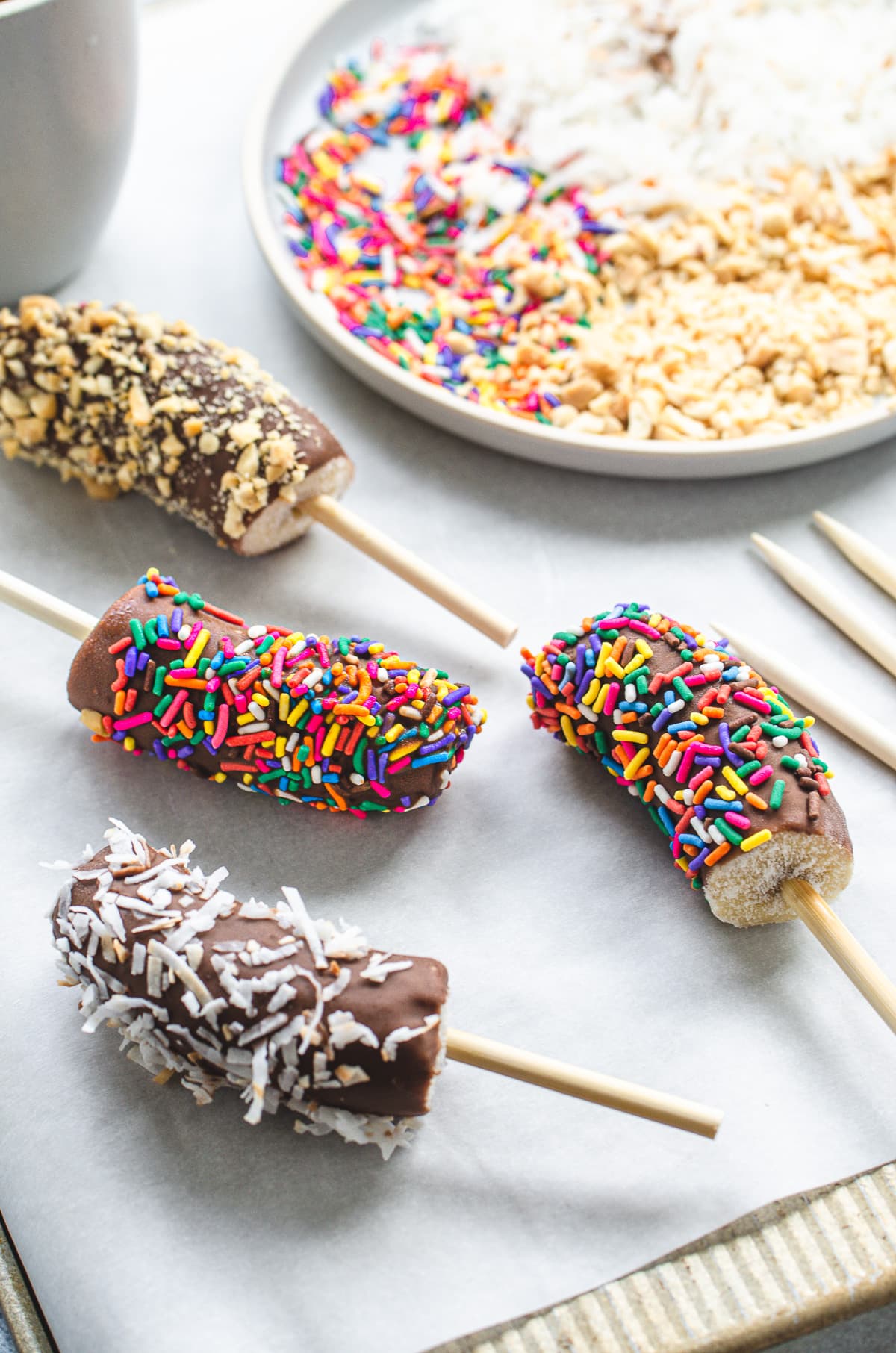 Chocolate dipped frozen banana pops on a sheet of parchment paper with a plate of sprinkle toppings in the background.