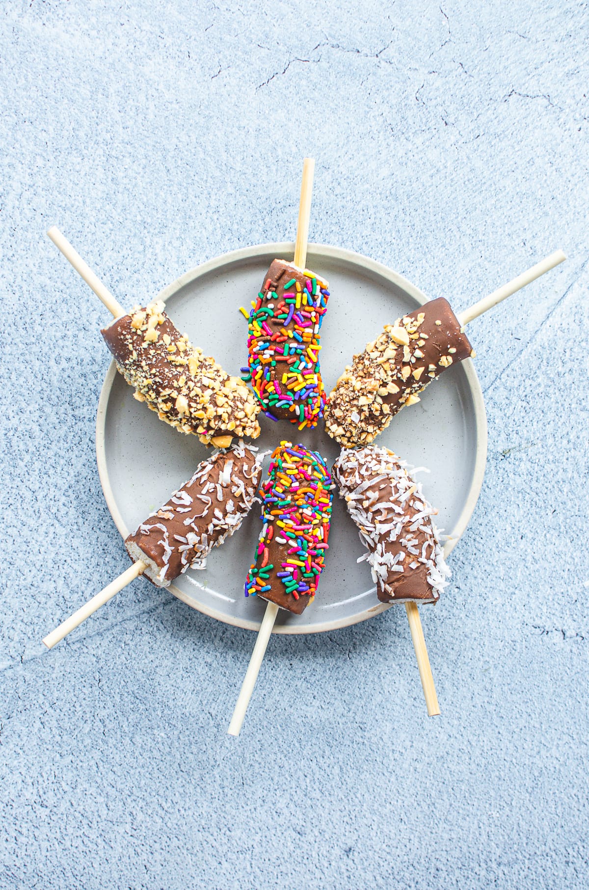 A blue plate with six chocolate-dipped frozen banana pops covered in an assortment of nuts, sprinkles, and coconut.