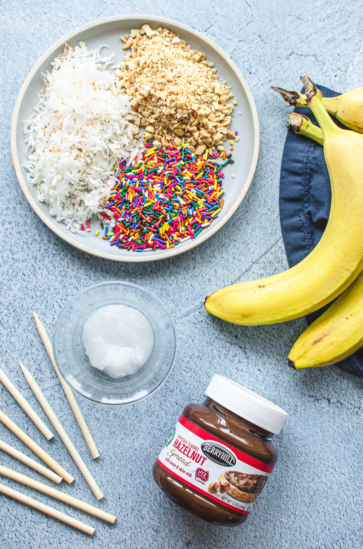 A plate of sprinkles, nuts and coconut flakes with bananas, Nutella, coconut oil, and wooden sticks on the side.