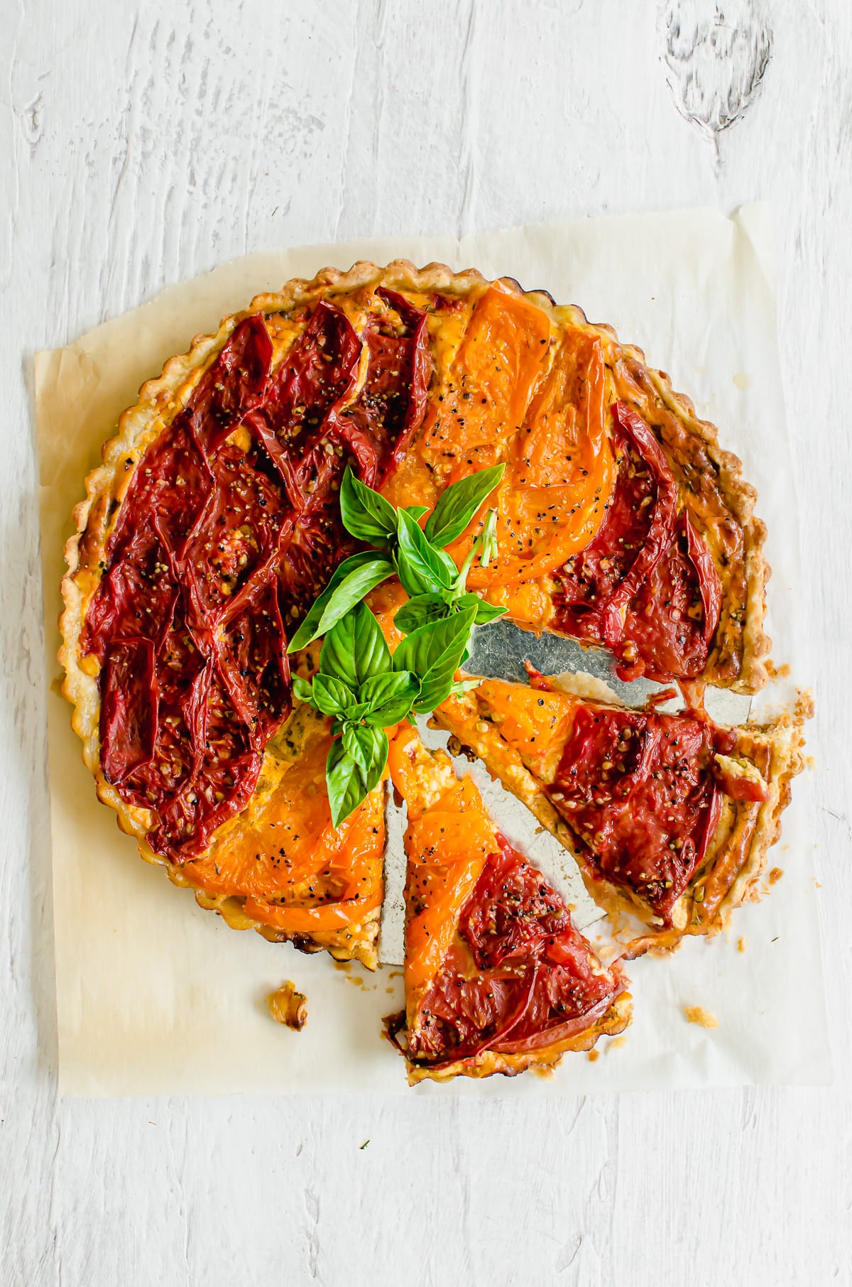 A tomato tart on parchment paper topped with basil sprigs and two slices of tart coming out on the side. 