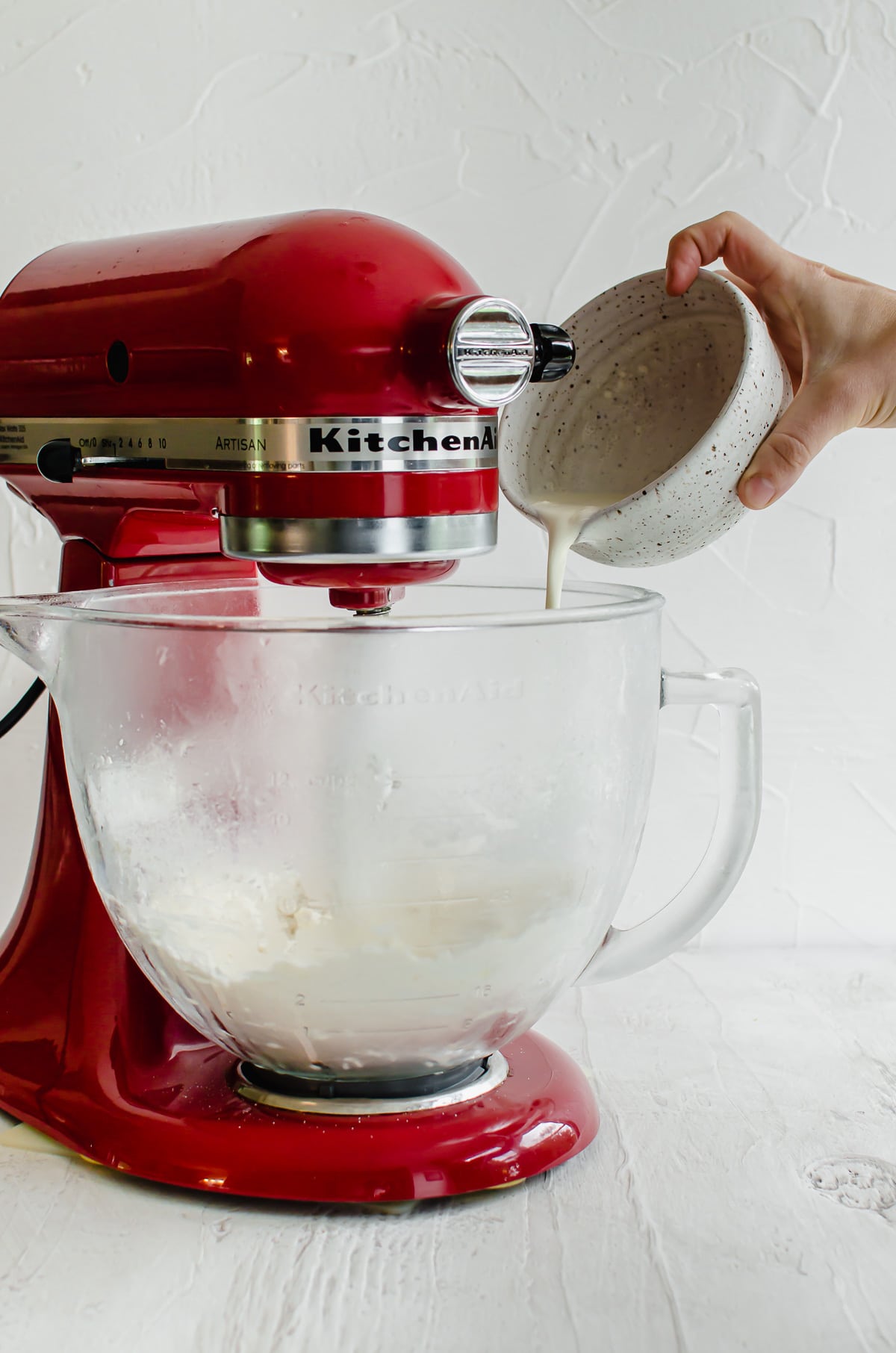 A hand pouring cream into a stand mixer bowl with dough inside. 