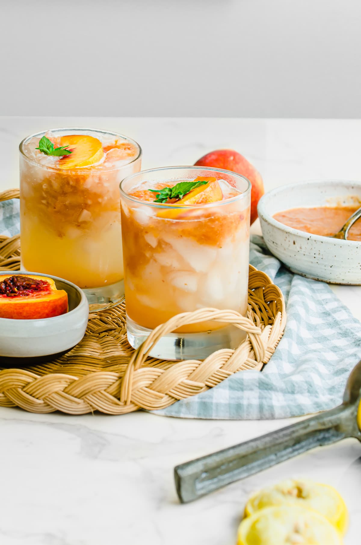 Two glasses of peach lemonade on a wicker serving tray with a gingham dish towel.