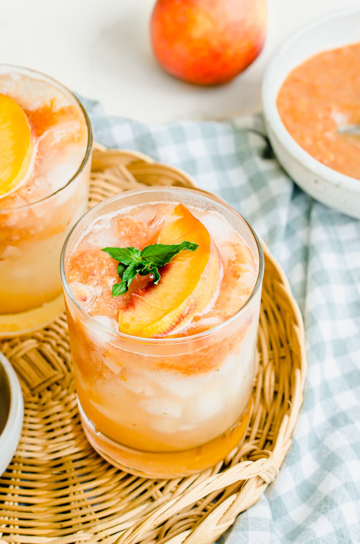 Two glasses of lemonade garnished with peaches on a wicker serving tray and a bowl of peach puree on the side.