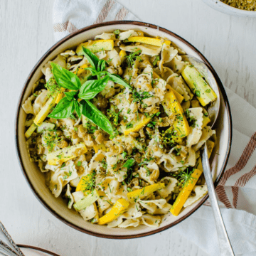 A brown-rimmed bowl with zucchini pasta salad and a spoon on the side.