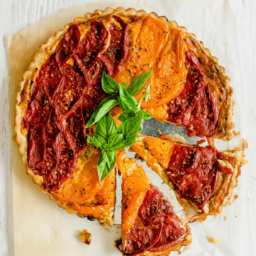 A tomato tart on parchment paper topped with basil sprigs and two slices of tart coming out on the side.