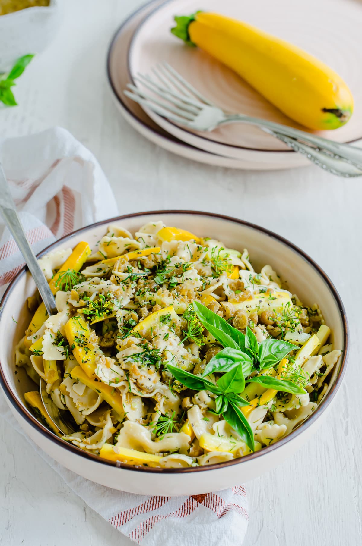 A bowl of summer pasta salad with plates and a zucchini in the background.