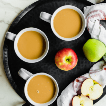 A black tray with three mugs of caramel apple cider and apples on the side.