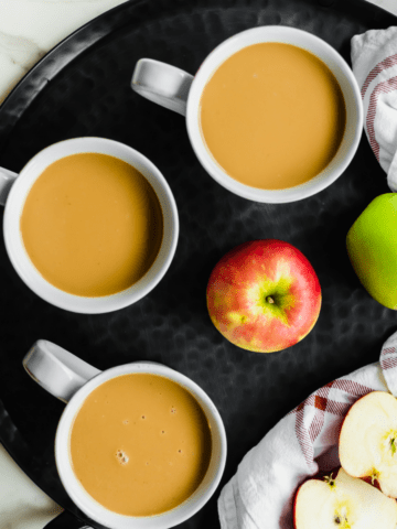A black tray with three mugs of caramel apple cider and apples on the side.