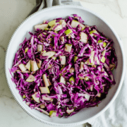 A white bowl filled with red cabbage apple slaw on a marble counter with a grey dish towel.