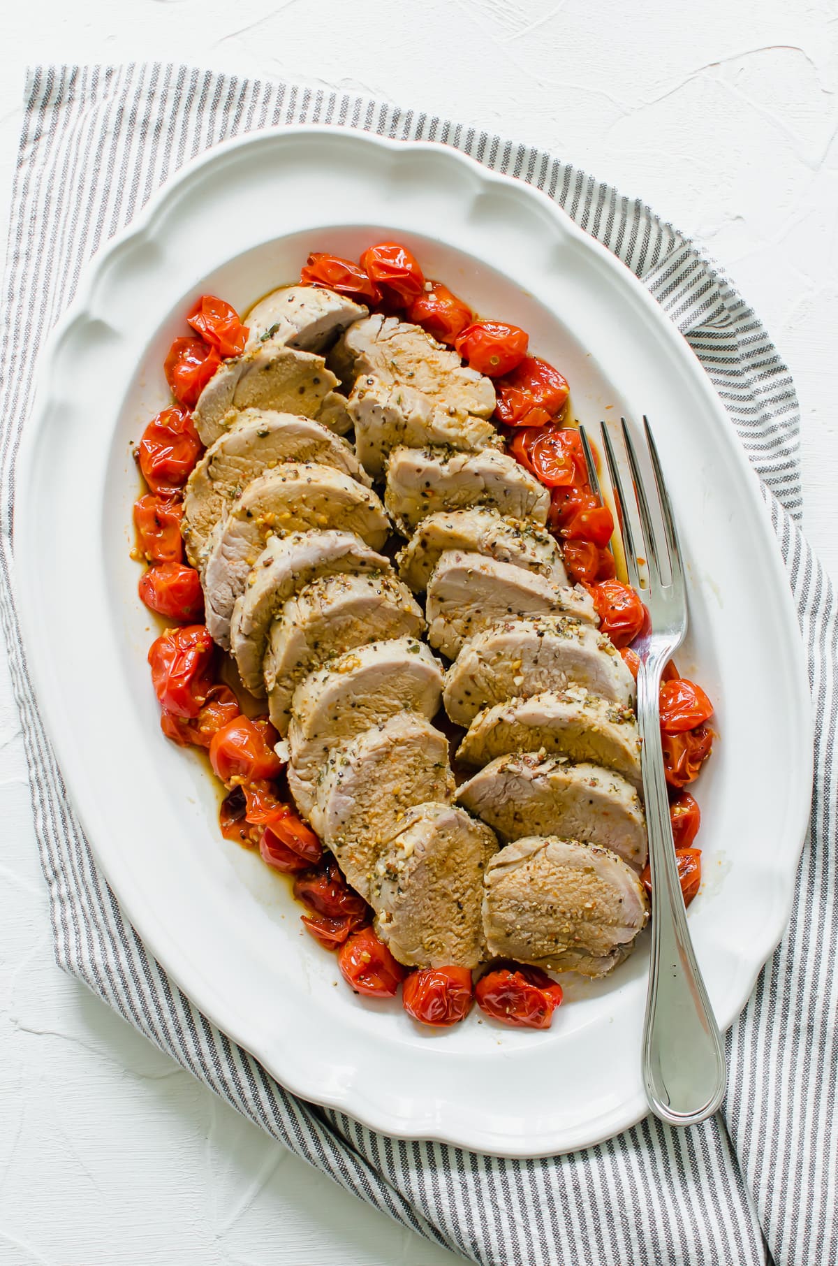 Slices of Tuscan roasted pork surrounded by roasted tomatoes on a white platter.