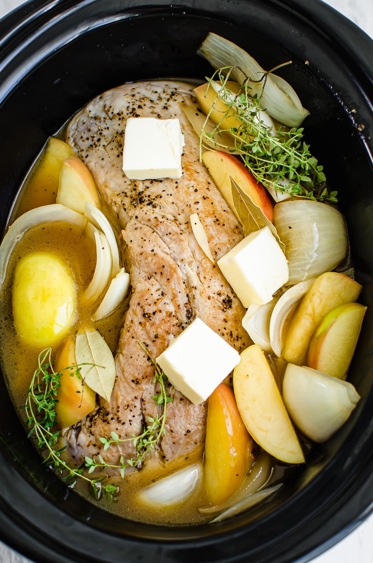 A pork loin roast in a slow cooker with apples, onions, thyme, butter pats, and apple cider.