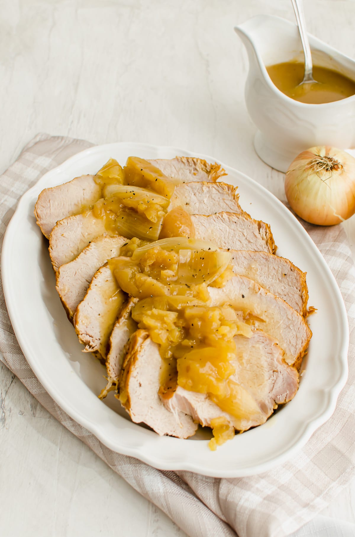 Slices of pork loin with apples and onions on a white platter with a gravy boat on the side.