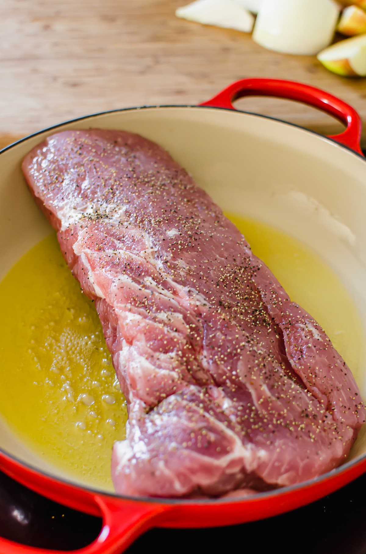 A pork loin roast being seared in olive oil in a red cast iron braiser pot.