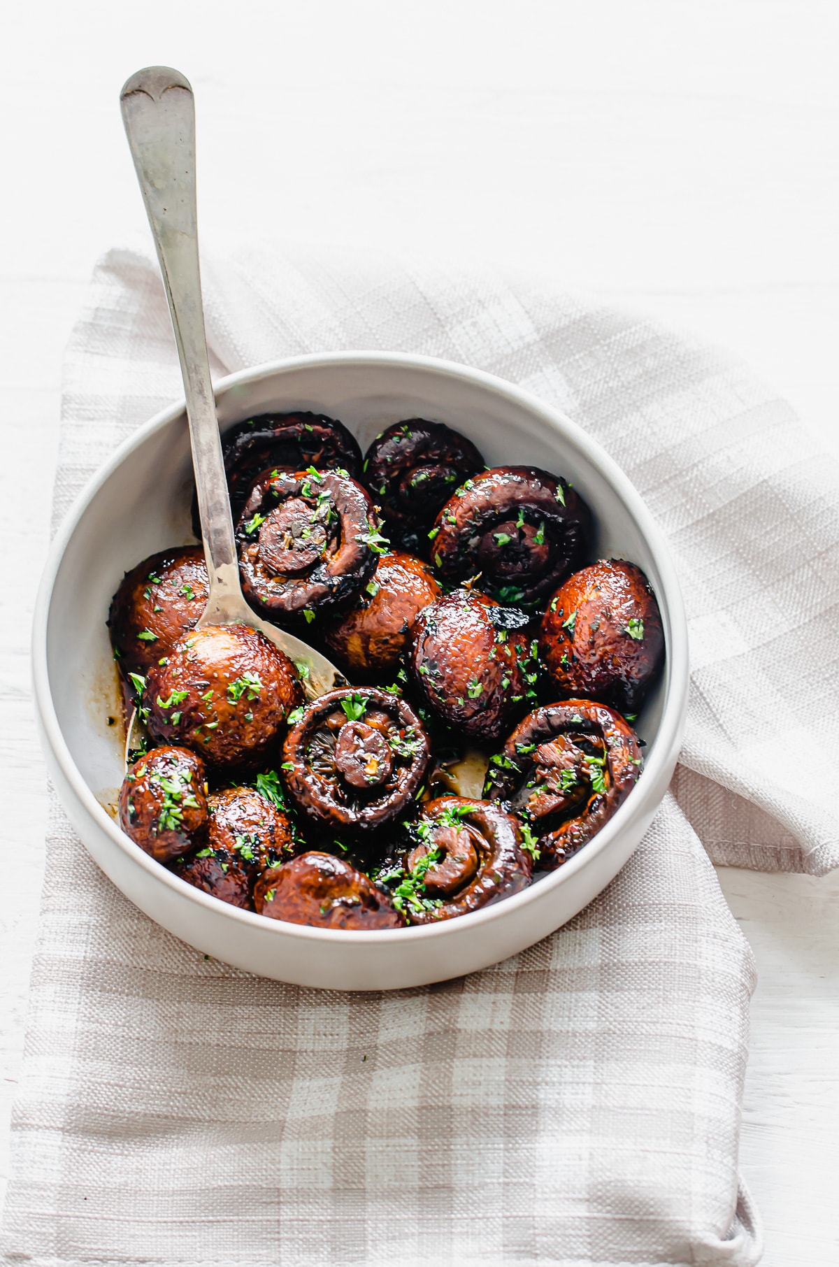 Overhead shot of a white serving bowl filled with roasted mushrooms on a beige plaid napkin.