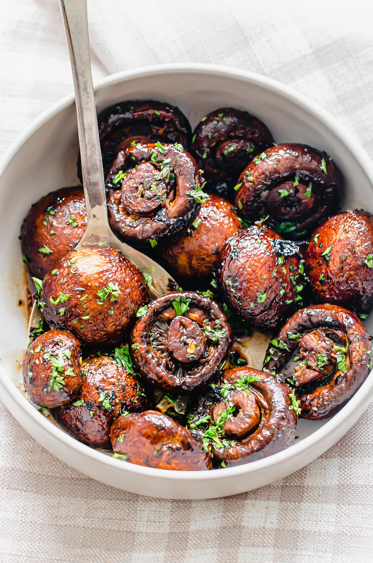 Roasted mushrooms garnished with parsley in a white bowl with a serving spoon.