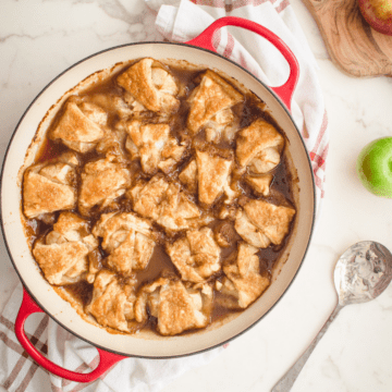 Apple dumplings in a round red baking dish with a serving spoon on the side.
