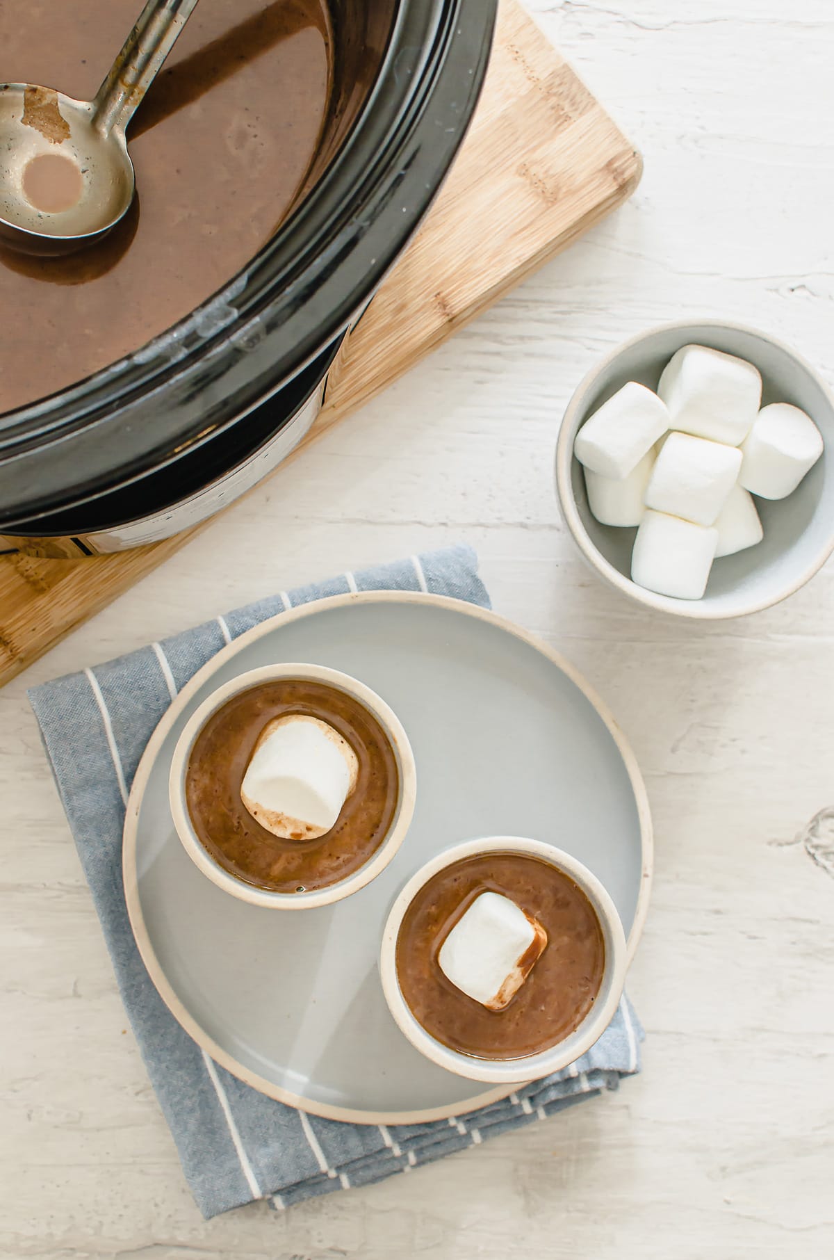 A slow cooker with hot chocolate next to two cups of chocolate with a bowl of marshmallows on the side.